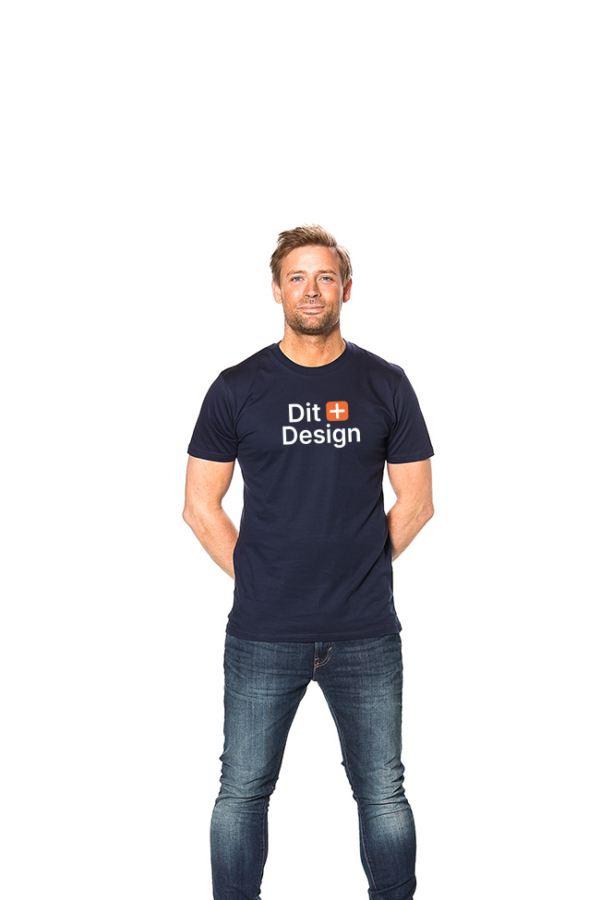 Mand i mørk t-shirt med teksten "Dit Design" står smilende med hænderne bag ryggen i en neutral, lys baggrund. Han bærer også jeans.