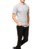 En mand med lyst hår iført en grå t-shirt, sorte bukser og blå sneakers smiler og står med hænderne i lommerne på en hvid baggrund.