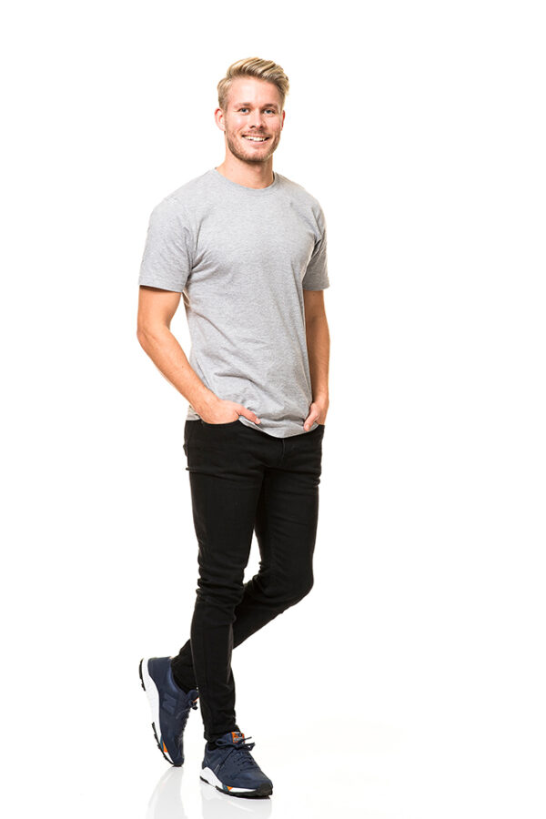 Heavy Lux T-shirt Økologisk En mand med lyst hår iført en grå t-shirt, sorte bukser og blå sneakers smiler og står med hænderne i lommerne på en hvid baggrund.