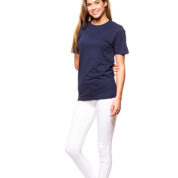 En kvinde iført en mørkeblå t-shirt og hvide bukser står smilende i en studieindstilling med hvid baggrund. Hun bærer sorte sneakers med lyserøde detaljer.