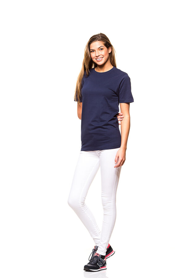 Heavy Lux T-shirt Økologisk navy dame En kvinde iført en mørkeblå t-shirt og hvide bukser står smilende i en studieindstilling med hvid baggrund. Hun bærer sorte sneakers med lyserøde detaljer.
