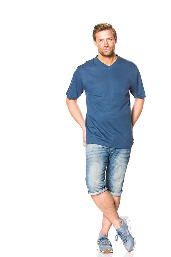 Klassisk V-hals Økologisk blå Mand iført en blå t-shirt og knælange denimshorts, står afslappet med krydsede ben og hænderne delvist i lommerne, på en hvid baggrund.