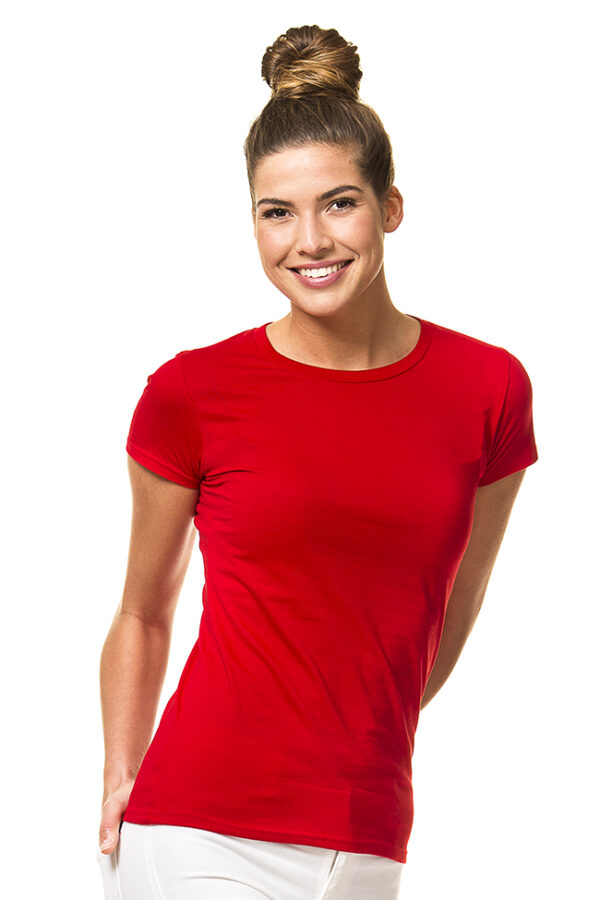 Lady Fitted Økologisk rød Kvinde iført rød t-shirt og hvid bukser, står og smiler med hænderne delvis i lommerne. Hendes hår er sat i en høj knold. Baggrunden er hvid.