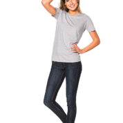 Kvinde iført grå t-shirt og mørke jeans står afslappet mod hvid baggrund, med den ene hånd på hoften og den anden hånd i håret.