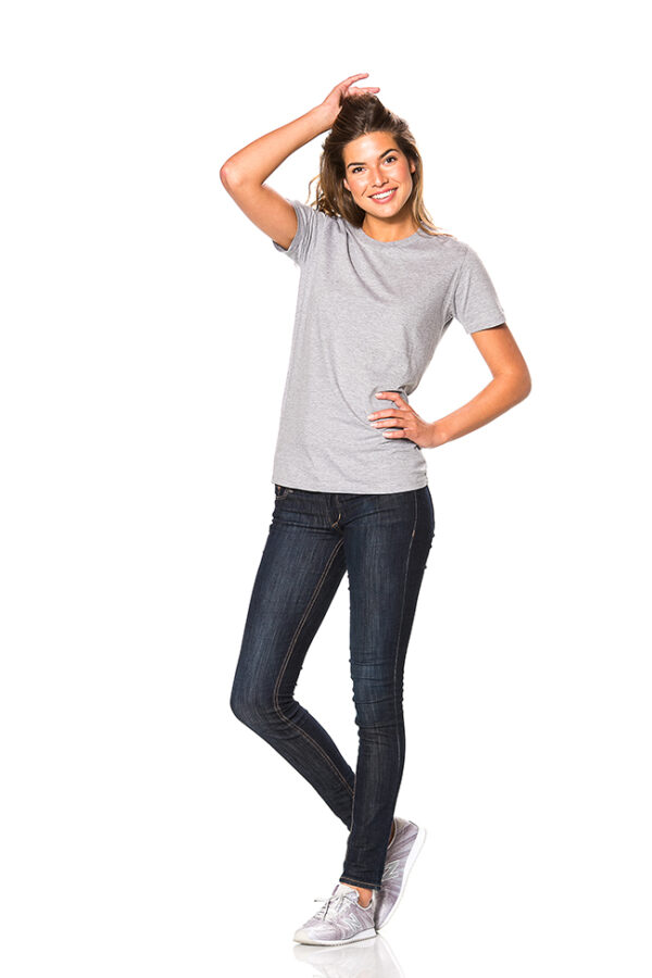 Mens Fitted Økologisk Kvinde iført grå t-shirt og mørke jeans står afslappet mod hvid baggrund, med den ene hånd på hoften og den anden hånd i håret.