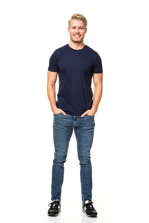 Mens Fitted Økologisk navy En mand står og smiler, iført en mørkeblå t-shirt, jeans og sorte sneakers, med hænderne i lommerne. Baggrunden er hvid og minimalistisk.