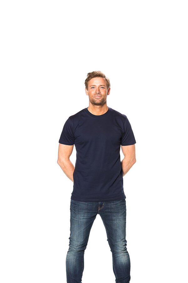 Mens Fitted Økologisk navya Mand står stille, iført mørkeblå t-shirt og jeans, med hænderne bag ryggen, mod hvid baggrund.