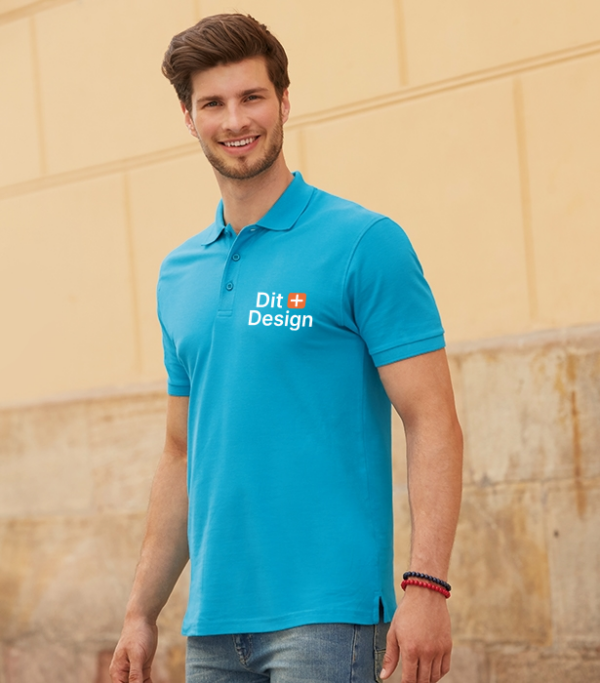 Mand iført en lyseblå polo med teksten "Dit Design" går foran en beige murstensvæg, med et venligt smil. Han bærer jeans og armbånd på håndleddet.