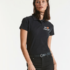 Person i sort polo-shirt står oprejst. Skjorten har teksten "Dit Design" ved brystet. Personen har sort hår og bærer et sort bælte. Baggrunden er lys.