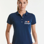 Kvinde står iført en mørkeblå poloshirt med teksten "Dit Design" trykt på venstre side. Hun står mod en lys baggrund, ser direkte fremad.