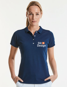 Kvinde står iført en mørkeblå poloshirt med teksten "Dit Design" trykt på venstre side. Hun står mod en lys baggrund, ser direkte fremad.