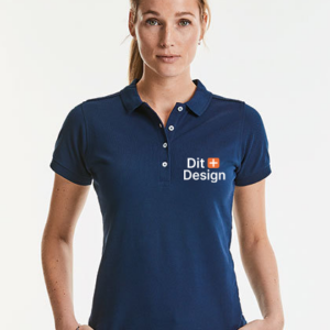 Kvinde står iført en mørkeblå poloshirt med teksten "Dit Design" trykt på venstre side. Hun står mod en lys baggrund, ser direkte fremad.
