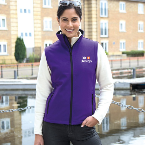 Kvinde står ved siden af vand i en bymiljø, iført en lilla vest med teksten "Dit Design." Baggrunden består af bygninger med vinduer og en række træer.