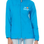 En person bærer en lyseblå jakke med lynlås, der har teksten "Dit Design" trykt i hvidt på brystet. Personen står mod en enkel lys baggrund.