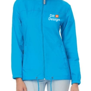 En person bærer en lyseblå jakke med lynlås, der har teksten "Dit Design" trykt i hvidt på brystet. Personen står mod en enkel lys baggrund.