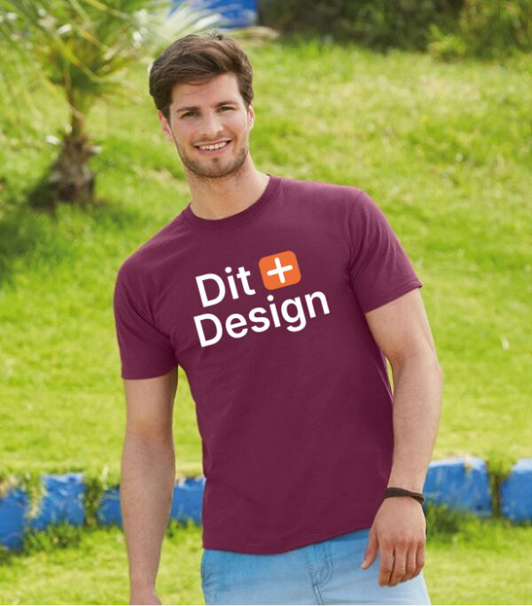 Mand iført bordeaux t-shirt med teksten "Dit Design" går i en græsklædt park. Der er en lav mur i blå og hvide farver i baggrunden.