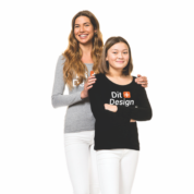 To kvinder står sammen; den ene holder den anden på skuldrene. De smiler i et lyst rum. Den ene har en sort bluse med teksten "Stay Unique".