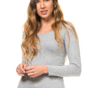 En kvinde med langt, bølget hår står i en grå trøje og hvide bukser, kigger direkte fremad og holder sine hænder sammen. Baggrunden er hvid.