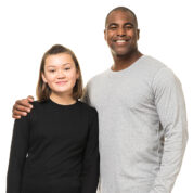 To personer står tæt sammen og smiler; en ung person i en sort trøje og en ældre person i en grå trøje. Baggrunden er hvid.