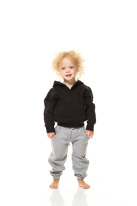 Et lille barn med krøllet hår står og smiler. Barnet har en sort hættetrøje og grå joggingbukser på. Baggrunden er hvid og enkel.