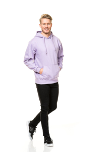En mand i en lilla hættetrøje og sorte bukser går og smiler. Han har hænderne i lommerne og bærer sorte sneakers. Baggrunden er helt hvid.