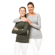 To kvinder står ved siden af hinanden i hvide bukser og langærmede bluser, hvor den ene har en grå top, og den anden har en mørkegrøn top. Hvid baggrund.