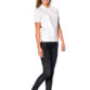 En kvinde står og smiler iført en hvid polo shirt, mørke jeans og sorte sneakers. Hun er i et studie og poserer mod en hvid baggrund.
