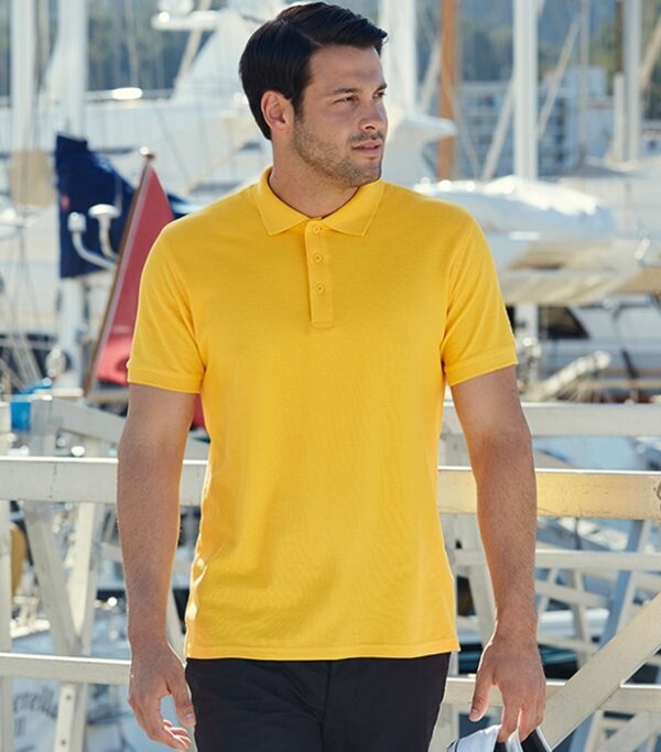 Premium Poloshirt Fruit gul En mand iført en gul polo t-shirt går udendørs på en bådebro. I baggrunden ses sejlskibe og master.