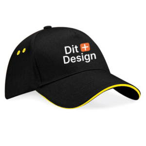 Sort kasket med teksten "Dit Design" og et orange plus-tegn på forsiden. Kasketten har gule detaljer på skyggen og ventilationshullerne.