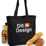 Sort mulepose med teksten "Dit Design" står oprejst. Ved siden af er der flasker med olie og brød, der skaber en indkøbs- eller picnicsammenhæng.