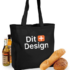 Sort mulepose med teksten "Dit Design" står oprejst. Ved siden af er der flasker med olie og brød, der skaber en indkøbs- eller picnicsammenhæng.