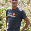 Manden smiler bredt, mens han står i en blomstrende have. Han har en grå T-shirt på med teksten "Dit + Design".