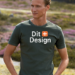 Mand står iført en grøn T-shirt med teksten "Dit Design" og et orange plus-symbol. Han står i et åbent, naturfyldt landskab med græs og marker i baggrunden.