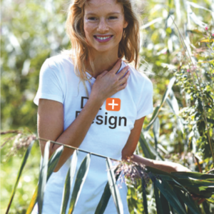 Kvinde smiler, iført en hvid T-shirt med teksten "D+ Design" i en frodig, grøn have.