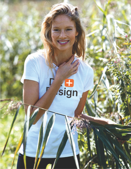 Kvinde smiler, iført en hvid T-shirt med teksten "D+ Design" i en frodig, grøn have.