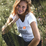 En person smiler og sidder i græsset iført en hvid T-shirt med teksten "Our Generation." Omkring hende er der vilde blomster og sollys.
