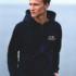 Mand iført mørk hættetrøje står ved vandkanten. Tekst på trøjen: "Dit Design." Han kigger til siden, mens det rolige vand udgør baggrunden.