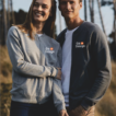 To personer står sammen iført grå trøjer med et logo. De smiler og hviler arme på hinanden. Baggrunden består af højt græs og træer i dagslys.