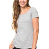 Kvinde smiler iført en grå T-shirt og sorte bukser, omgivet af en hvid baggrund. Hun har langt, bølget hår og står med hænderne i lommerne.