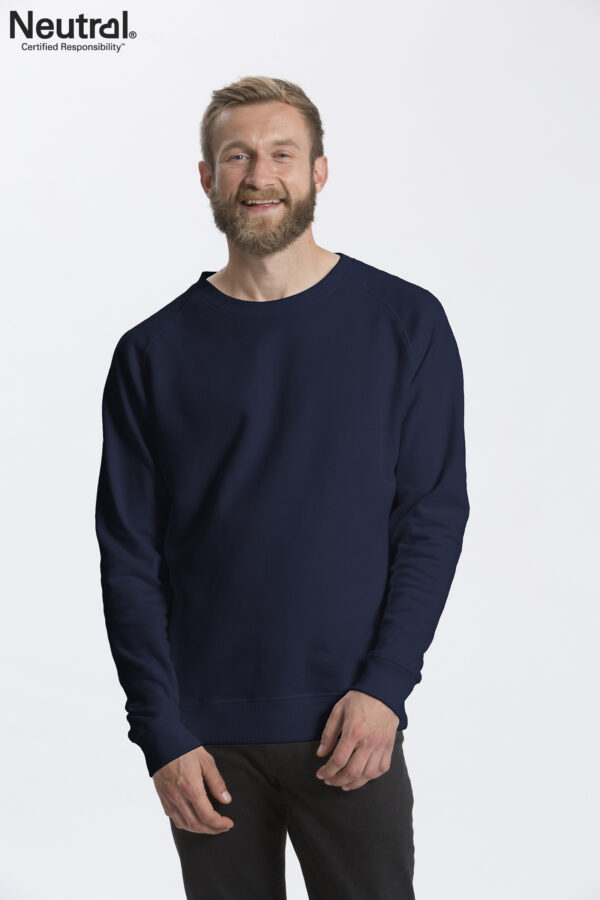 O63001_M_NAV En mand med skæg og kort hår smiler, iført en mørkeblå sweatshirt og sorte bukser. I øverste venstre hjørne er teksten: "Neutral® Certified Responsibility™." Baggrunden er hvid.