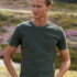 En mand iført en mørkegrøn t-shirt står med hænderne i lommerne i et grønt landskab med lilla blomster i baggrunden.