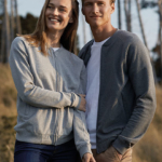 To personer, iført grå trøjer, står tæt sammen og smiler udendørs i en skov med høje træer.