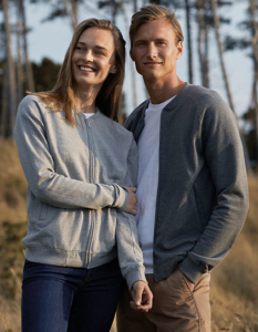 To personer, iført grå trøjer, står tæt sammen og smiler udendørs i en skov med høje træer.
