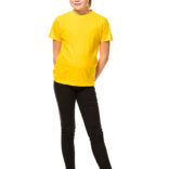 En pige iført en gul t-shirt og sorte bukser står afslappet med hænderne i lommerne og venstre ben krydset foran højre. Baggrunden er hvid og minimalistisk.