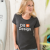 Kvinde står og smiler iført en sort T-shirt med teksten "Dit Design." Hun befinder sig i et lyst rum ved en surfbræt med skrift.