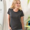 Kvinde iført mørkegrå t-shirt står afslappet ved siden af lysebrædder og surfbræt mod en trævæg.