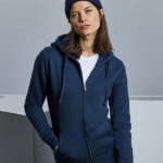 En person iført en navy blå hættetrøje og hue står afslappet med hænderne i lommerne i et minimalistisk, gråt rum.