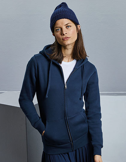 En person iført en navy blå hættetrøje og hue står afslappet med hænderne i lommerne i et minimalistisk, gråt rum.