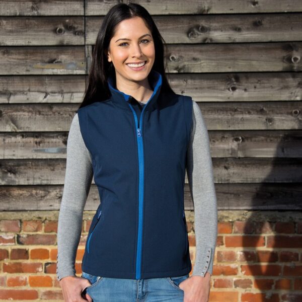 Klassisk damesoftshell vest stil En person står stille iført en blå vest og grå trøje. I baggrunden ses en væg af træplanker og mursten. Ingen tekst er synlig.