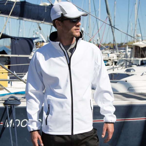 Klassisk herresoftshell hvid Mand iført hvid jakke og kasket står på en marina omgivet af både, kigger til siden med hånden i lommen.